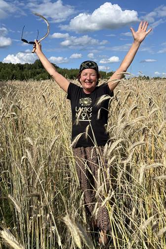 Margarita Macijauskienė stovi rugių lauke, pasiruošusi dalintis tradiciniais amatų įgūdžiais šiaudinių sodų rišimo dirbtuvėse.